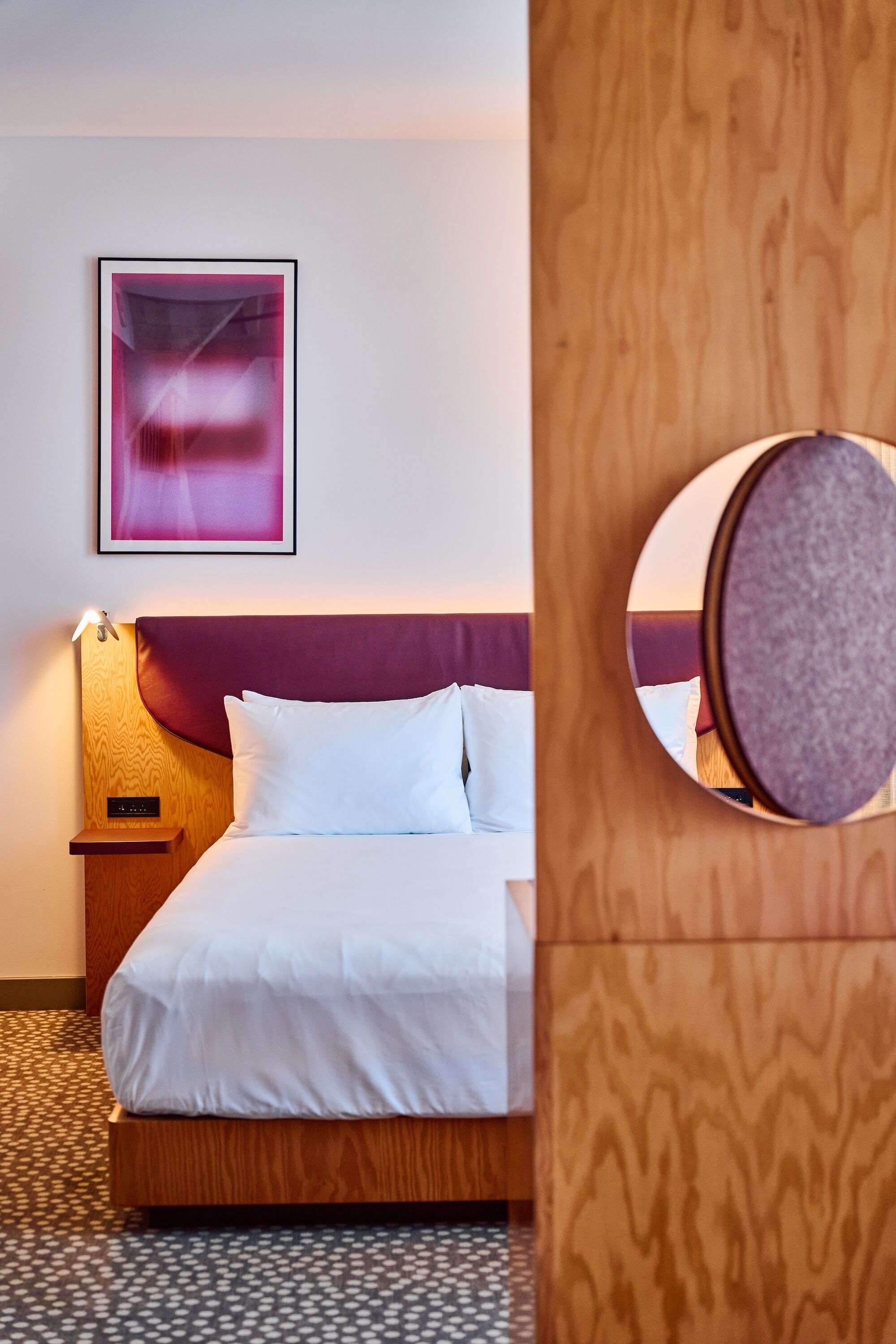 A hotel room with a bed, a nightstand, a wall-mounted picture, and a round mirror.