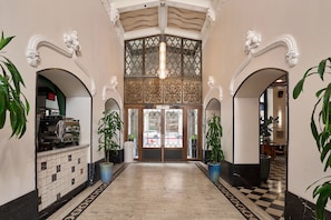 A hotel lobby with ornate architecture, a checkered floor, and potted plants.
