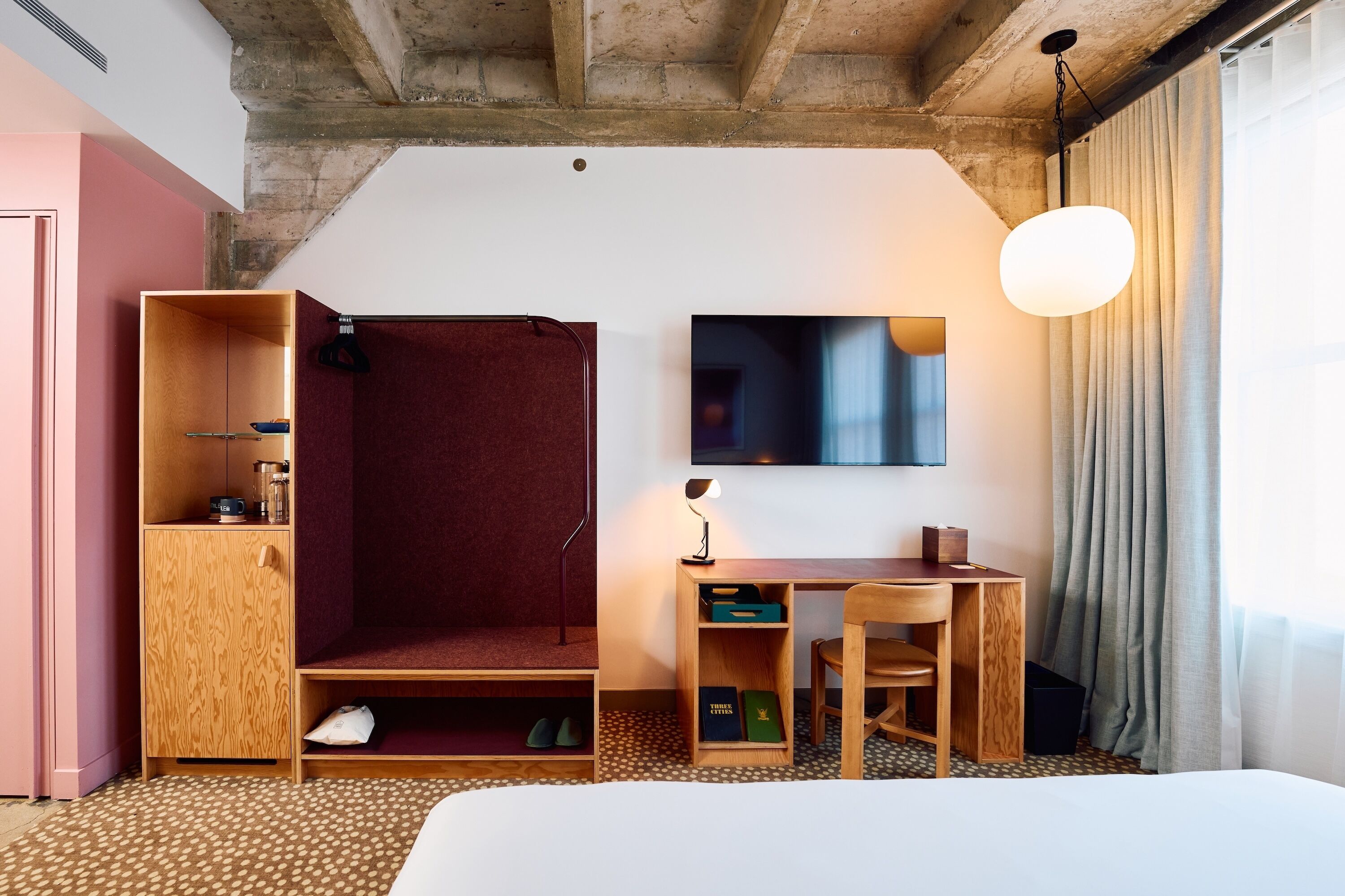 Hotellihuone, jossa on sänky, työpöytä tuolilla, vaatekaappi, televisio ja riippuvalaisin.