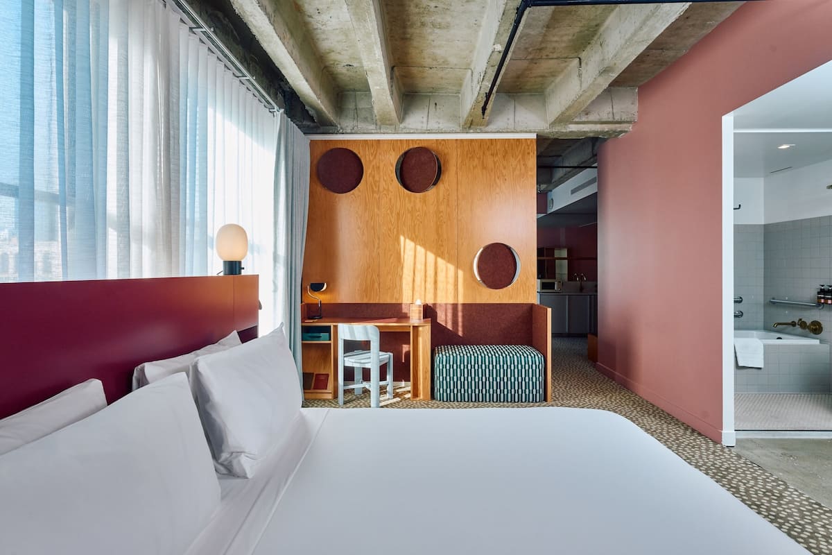 Hotellihuone, jossa on suuri sänky, työpöytä, tuoli, pieni pöytä, pyöreä peili ja kylpyhuone, joka näkyy avoimesta ovesta.