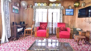 A wooden room with two red armchairs, a glass coffee table, a chandelier, a TV, and a patterned carpet. - Houseboat Zaindari Palace (Srinagar)