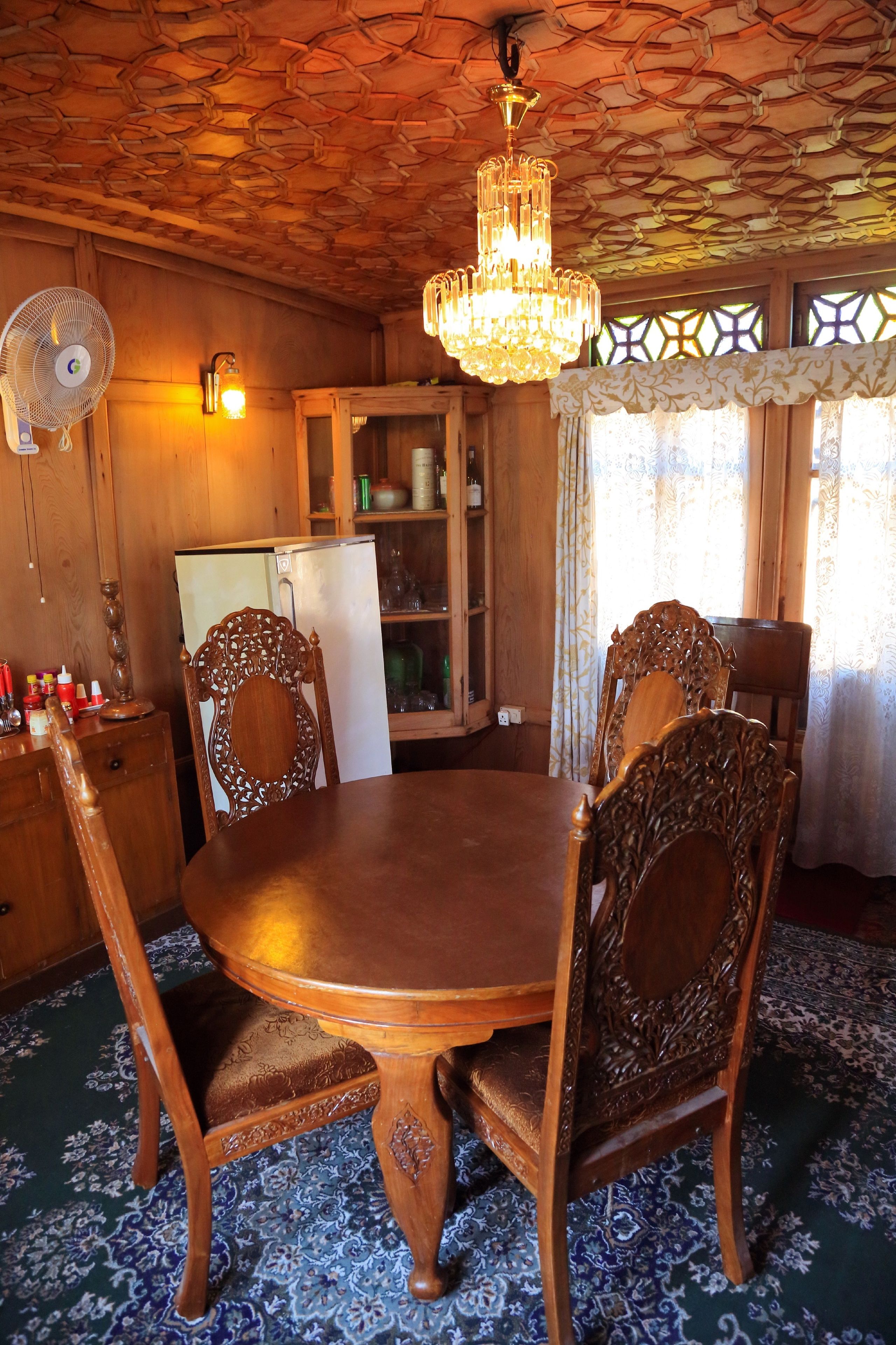 Uma sala de jantar de madeira com uma mesa redonda e cadeiras ornamentadas, um lustre e uma geladeira ao fundo.