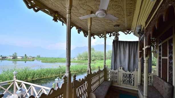 A covered porch with ornate railings, a ceiling fan, and a view of a lake and mountains. - Houseboat Zaindari Palace (Srinagar)