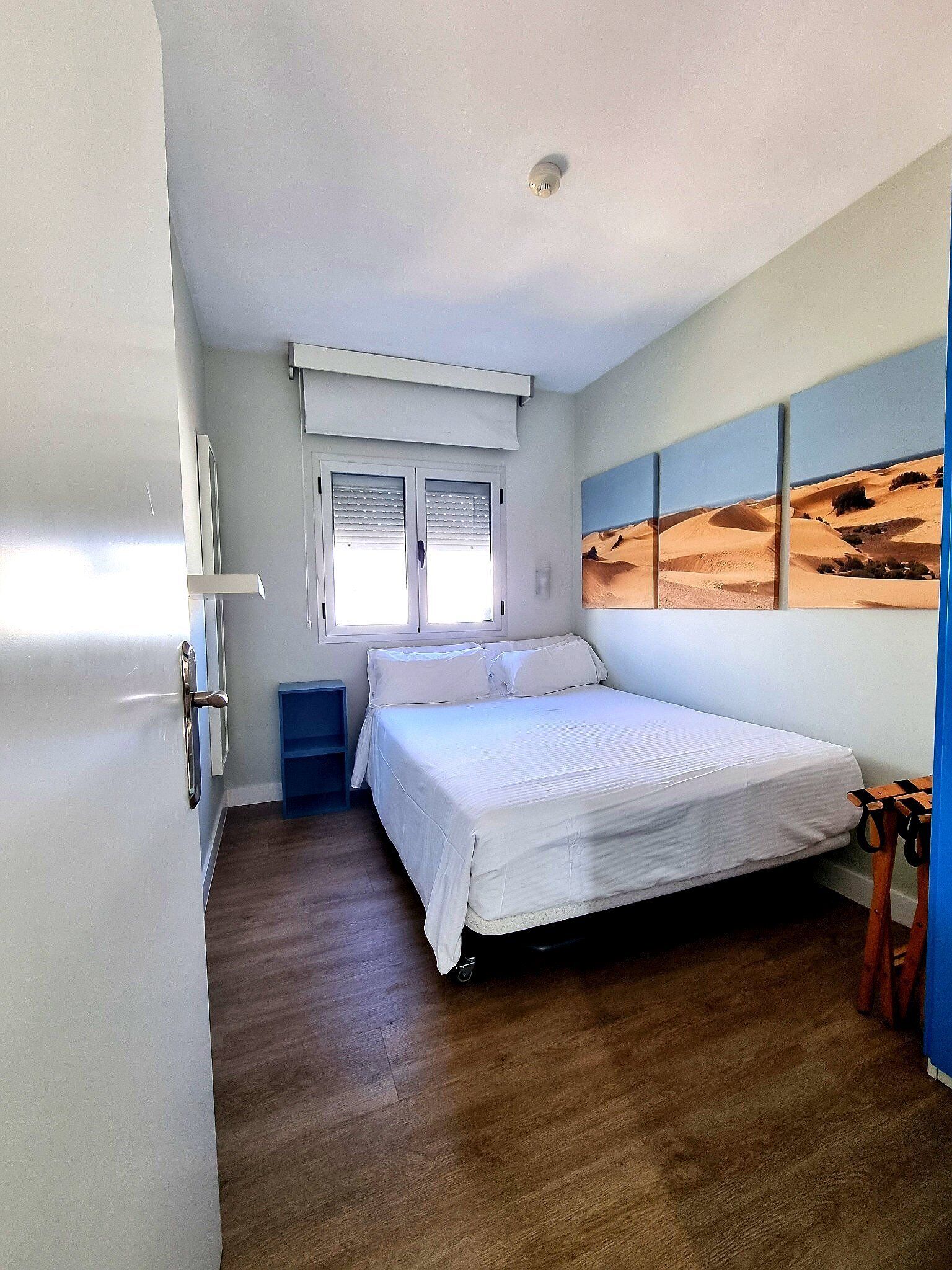 A bedroom with a bed, a window with blinds, a wall-mounted air conditioning unit, and a desert landscape painting.