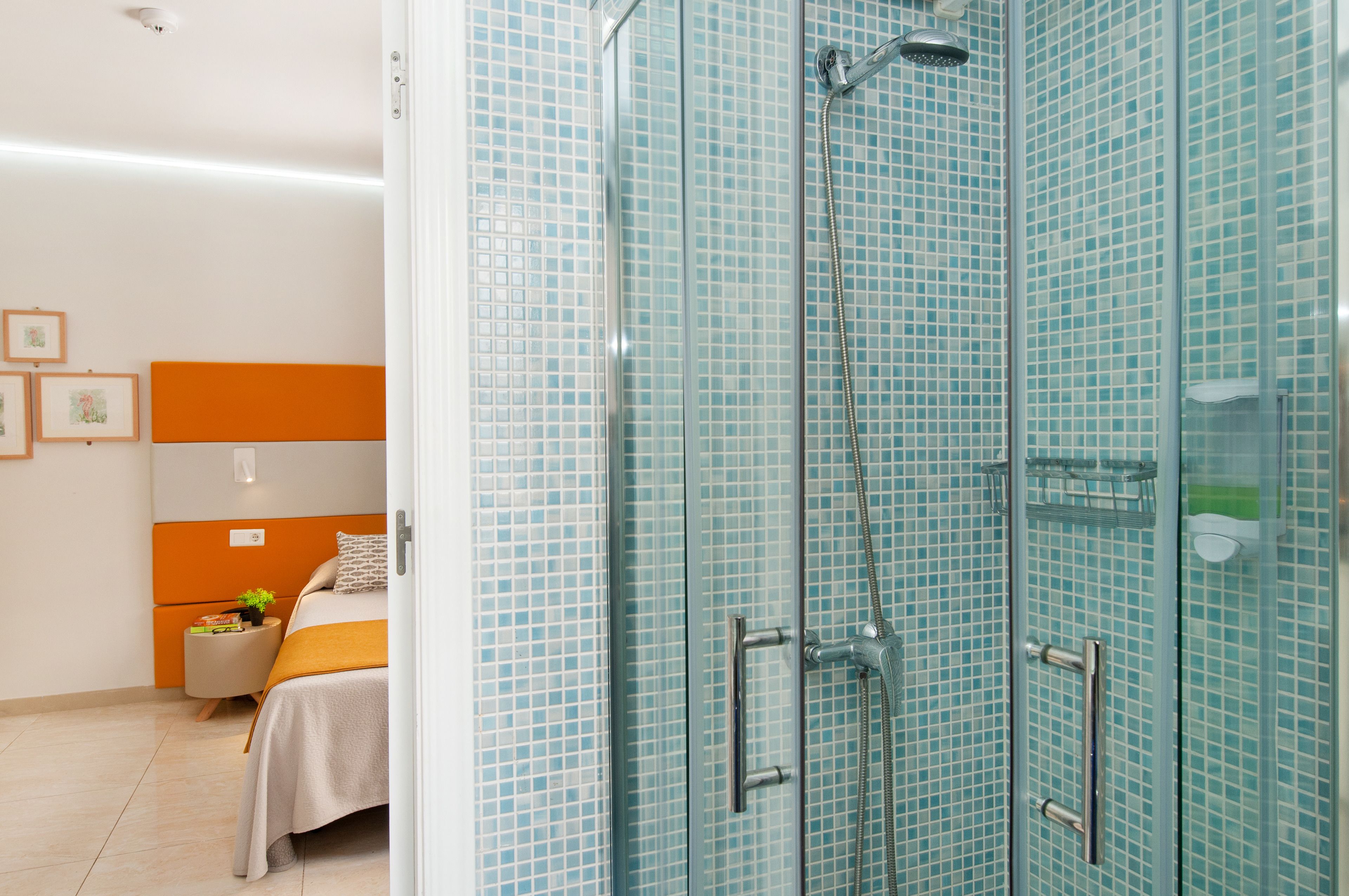 A modern bathroom with a glass shower enclosure, a bed with an orange headboard, and a small bedside table with a plant.