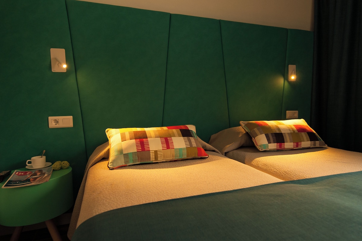 Une chambre d’hôtel avec deux lits, des murs verts et une petite table de chevet sur laquelle se trouvent un magazine et une tasse.