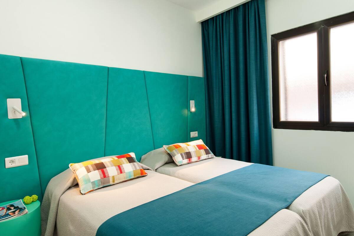 Ein Hotelzimmer mit türkisfarbenen Wänden, zwei Betten mit bunten Kissen, einem Fenster mit Vorhängen und einem kleinen Tisch mit einer Zeitschrift.