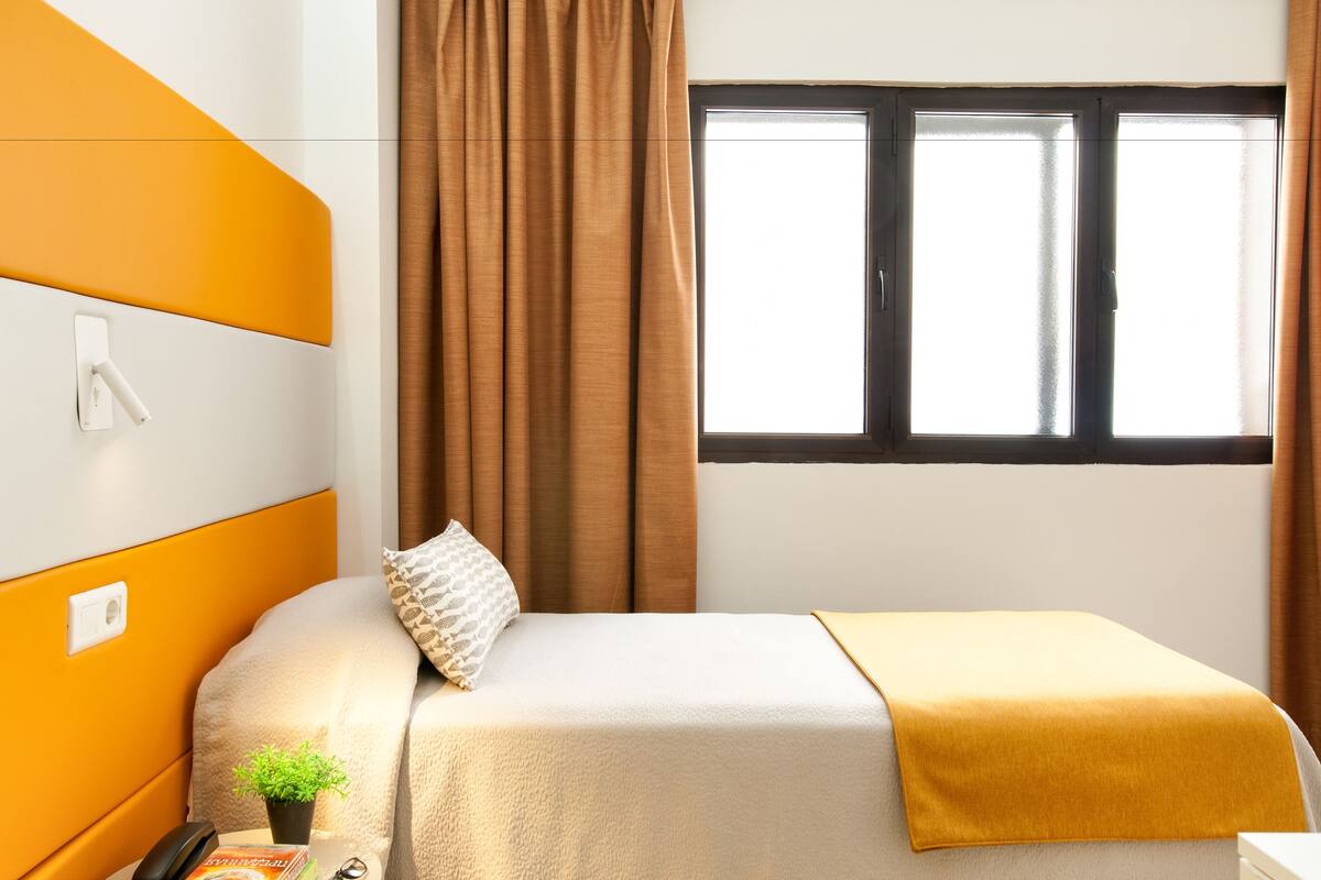 Ein ordentlich bezogenes Bett mit weißer und gelber Decke, einer kleinen Pflanze und einem Kissen an einem Kopfteil mit einer Steckdose.