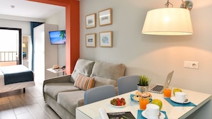 A modern hotel room with a sofa, a dining table with breakfast, a laptop, and a TV on the wall. - Hotel Apartamentos Bajamar (Las Palmas de Gran Canaria)