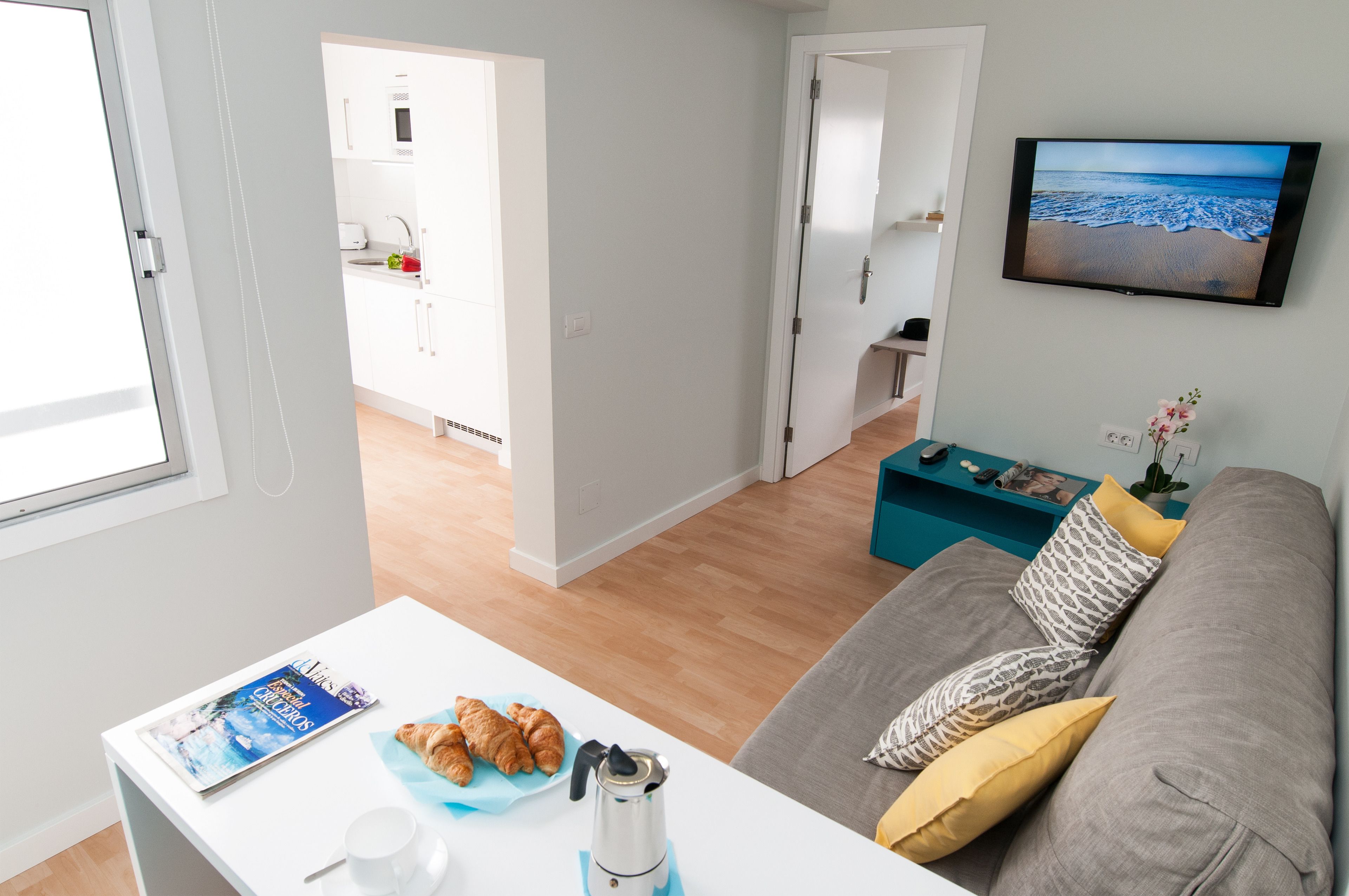 Un moderno salón con un sofá gris, una mesa de centro con cruasanes, una revista y una cafetera. Un televisor montado en la pared muestra una escena de playa.