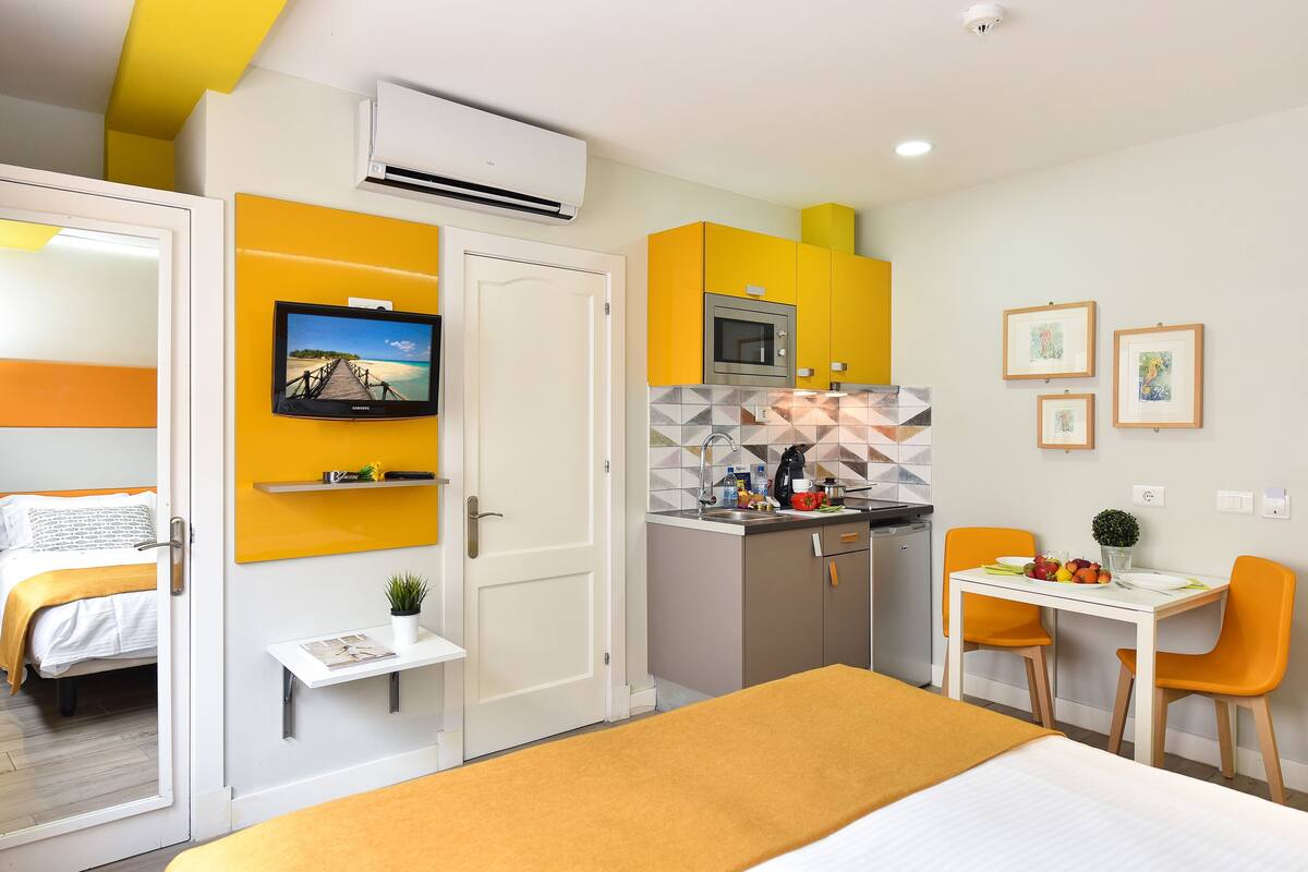 Ein kompaktes Hotelzimmer mit Bett, einer kleinen Küchenzeile, einem Essbereich und einem an der Wand befestigten Fernseher.