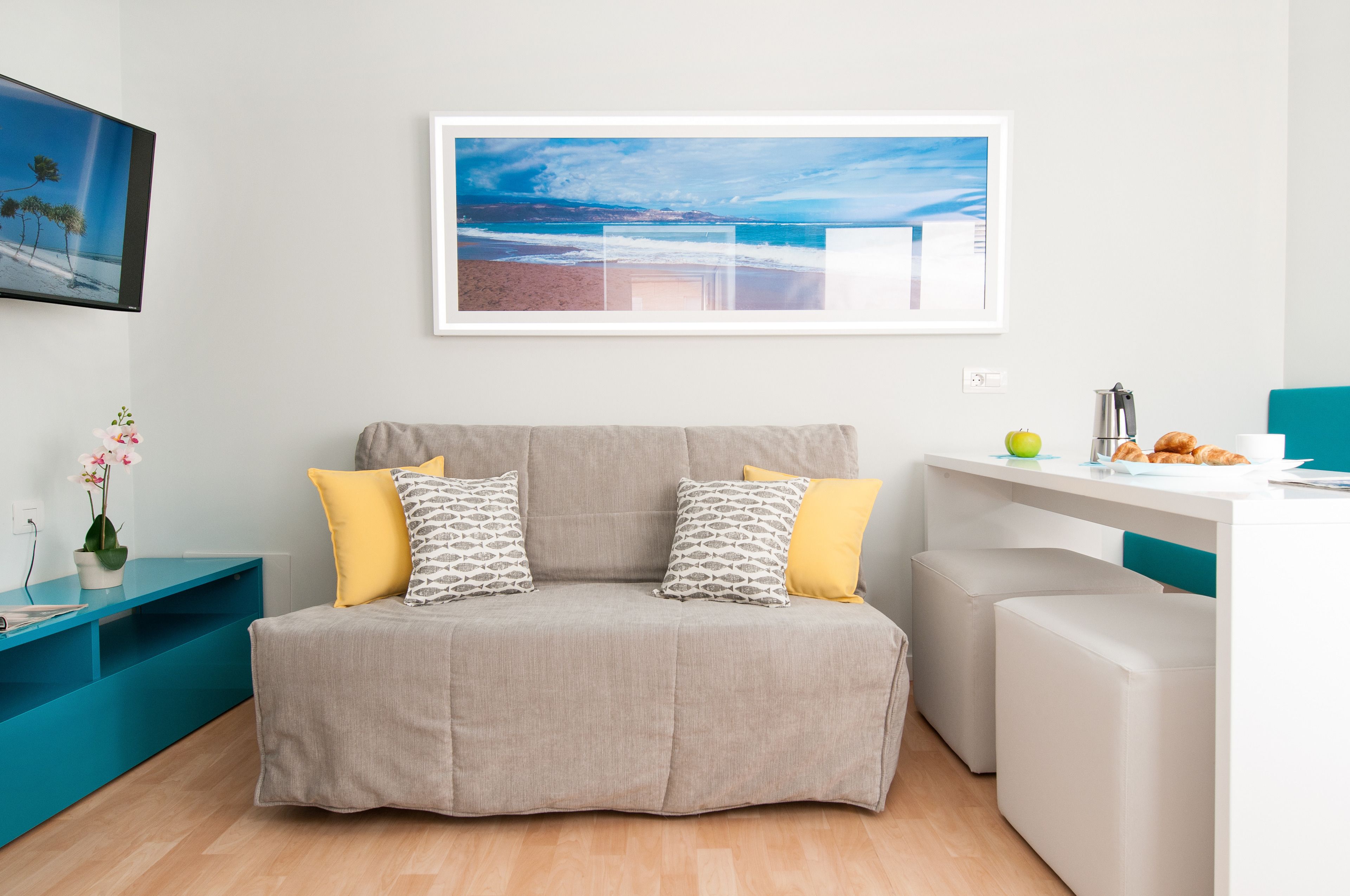 Una sala moderna con un sofá gris, cojines amarillos y estampados, una mesita de noche azul verdoso con una planta en maceta, y una escena de playa enmarcada en la pared.