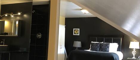 A modern hotel room with a large bed, a separate bathroom, a vanity with a mirror, and a small bedside table with a lamp.