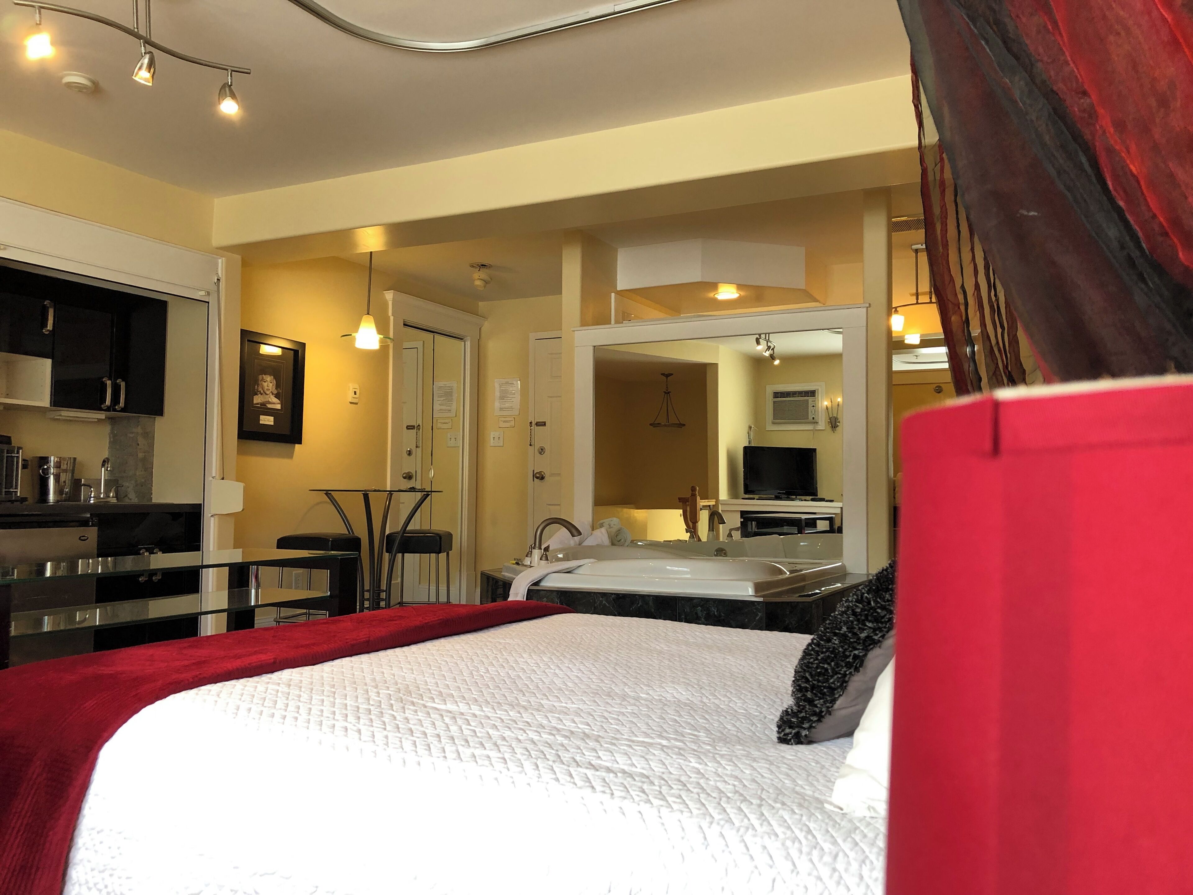 A bedroom with a bed, a desk, a chair, a TV, and a bathroom with a sink and mirror.