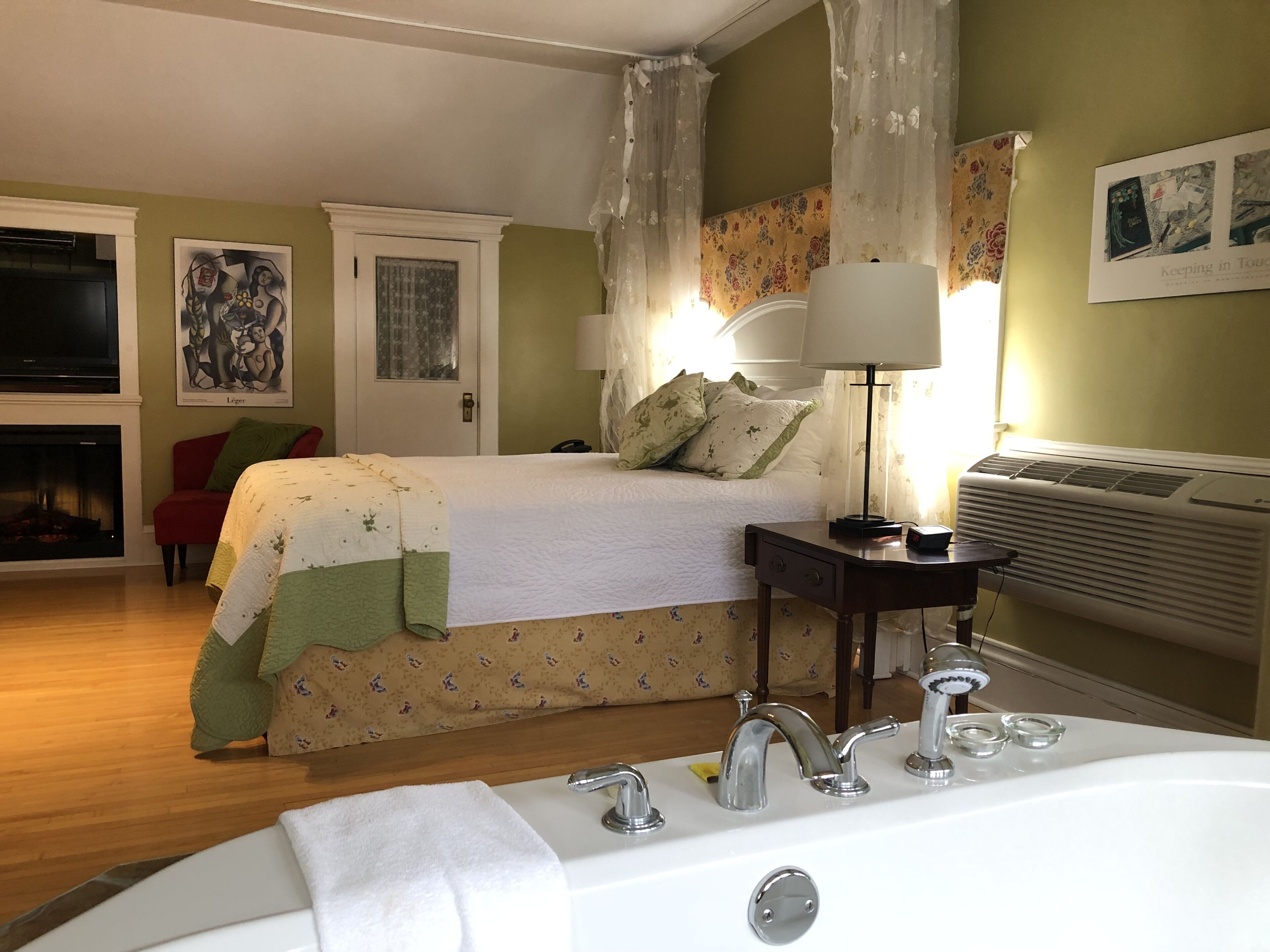 Habitación de hotel con cama, mesita de noche con lámpara, baño con bañera, chimenea y televisor.