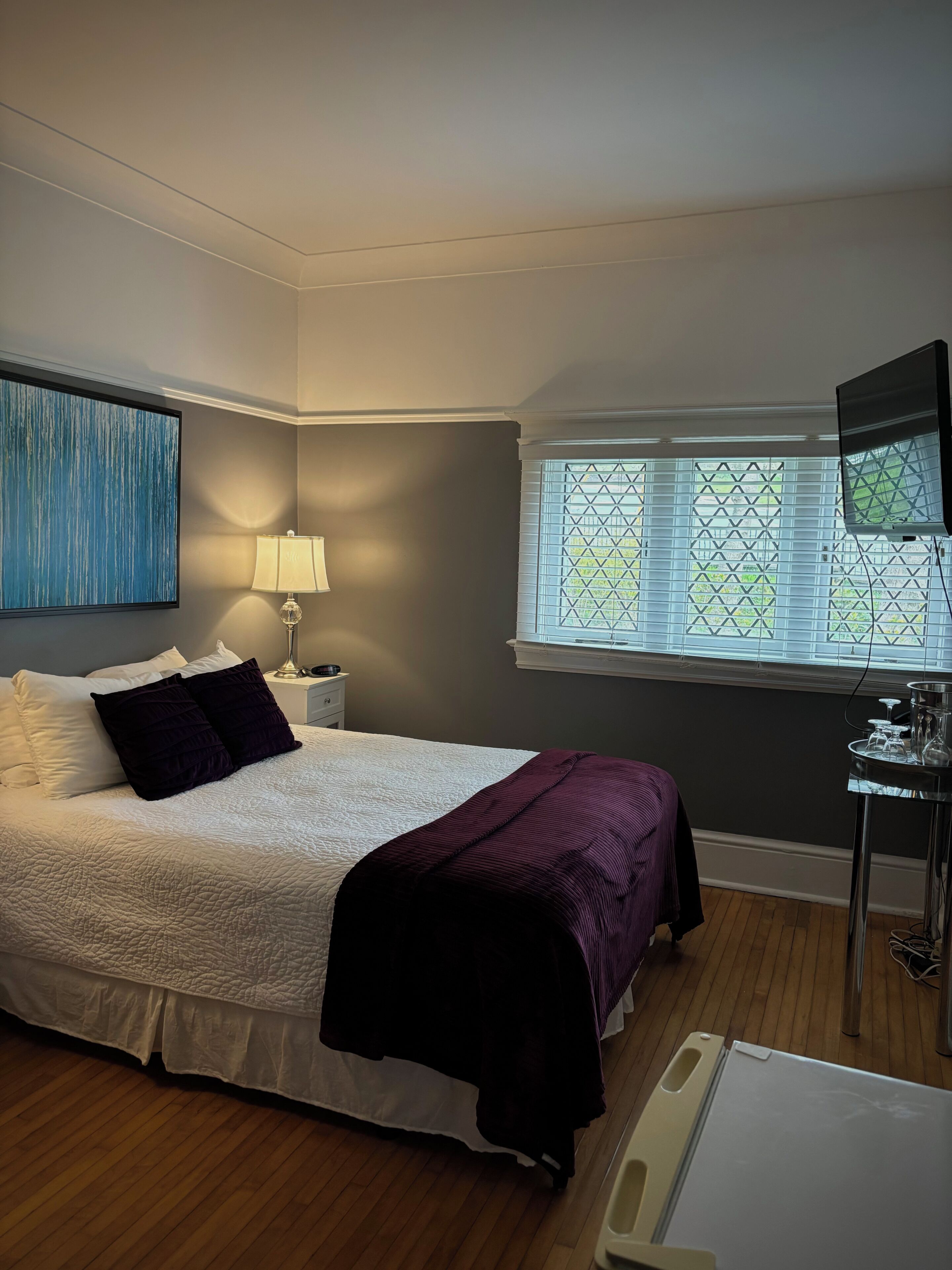 A bedroom with a bed, a nightstand, a lamp, a window with blinds, and a television.