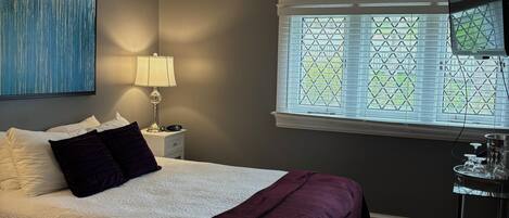 A bedroom with a bed, a nightstand, a lamp, a window with blinds, and a television.
