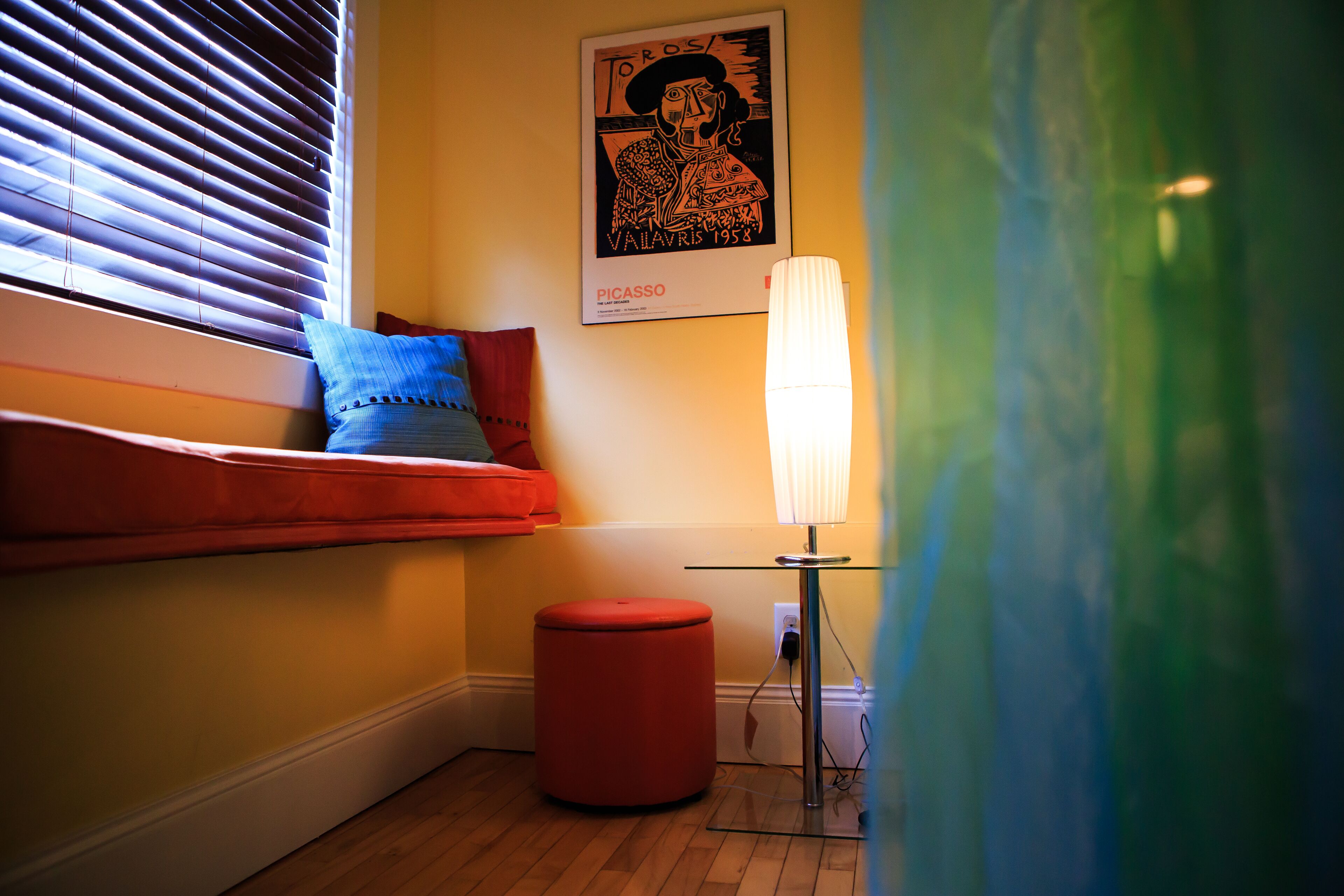 Una habitación con ventana, un reposapiés rojo, una lámpara y un cartel de Picasso en la pared.
