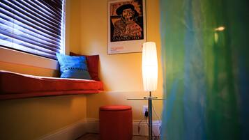 Una habitación con ventana, un reposapiés rojo, una lámpara y un cartel de Picasso en la pared.