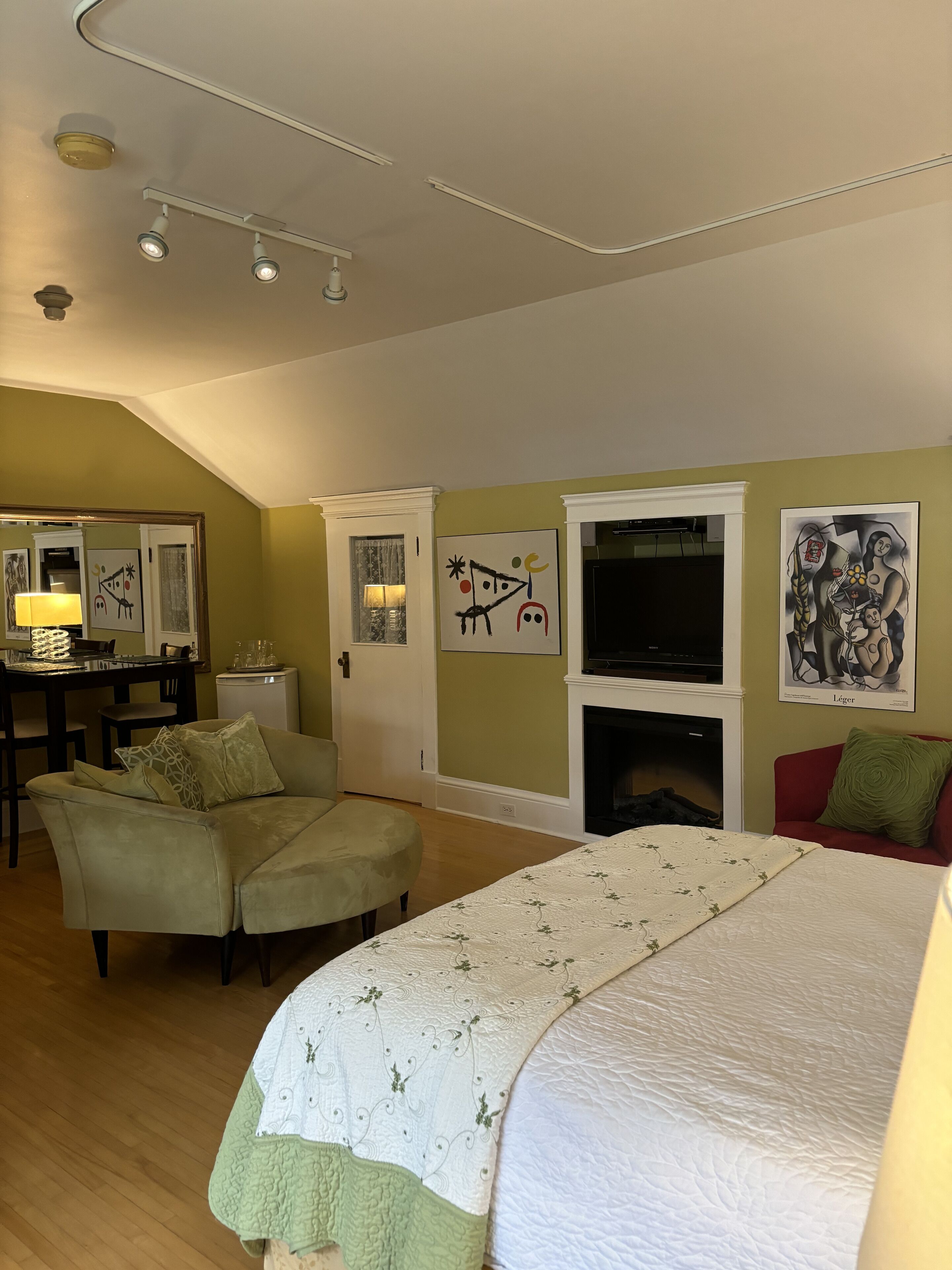Un dormitorio con una cama, dos sillas, un televisor, obras de arte en las paredes y una chimenea.