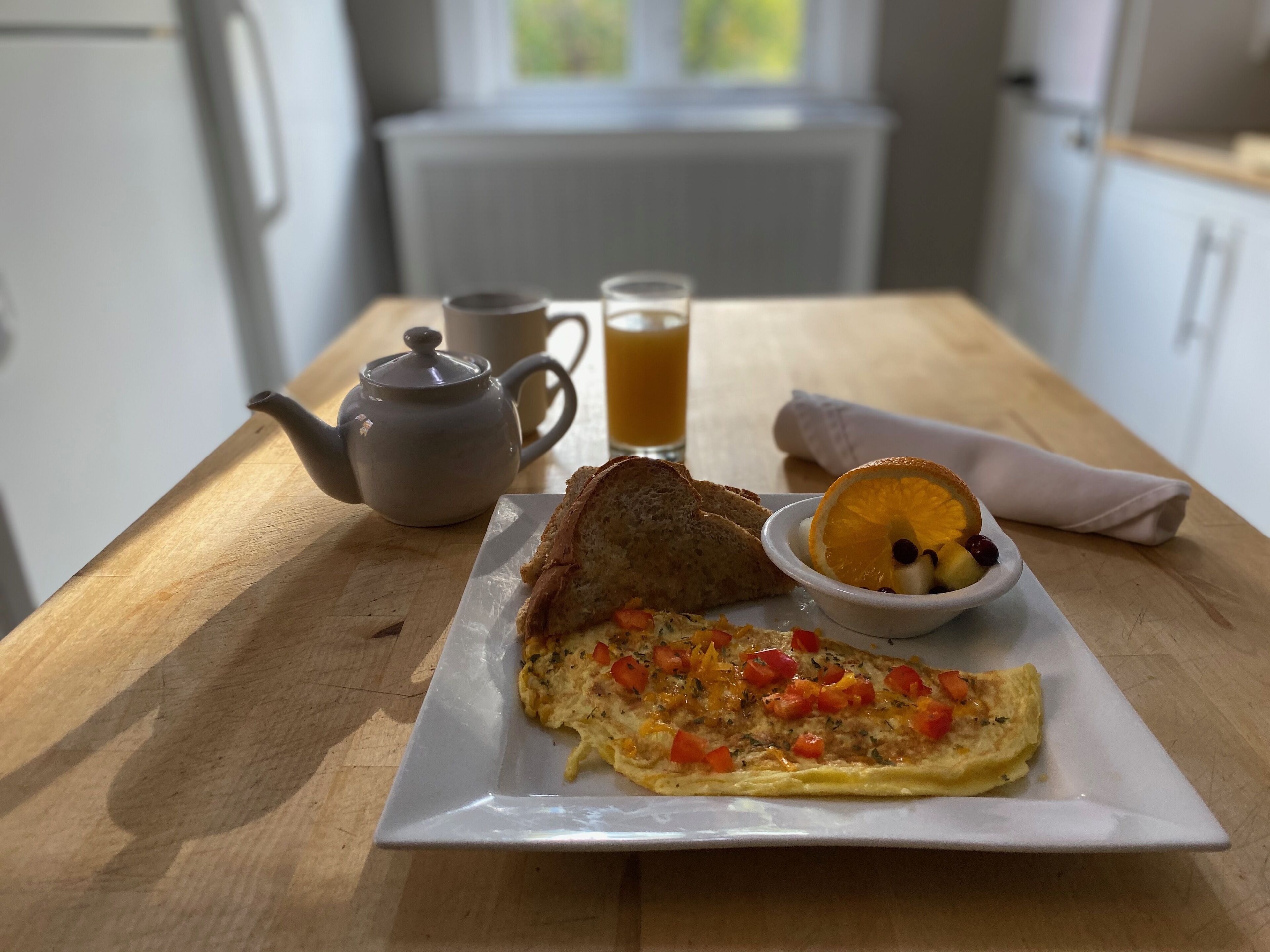 Une table en bois dressée pour le petit-déjeuner, avec des toasts, une omelette, une théière, un verre de jus d’orange et un bol de fruits.