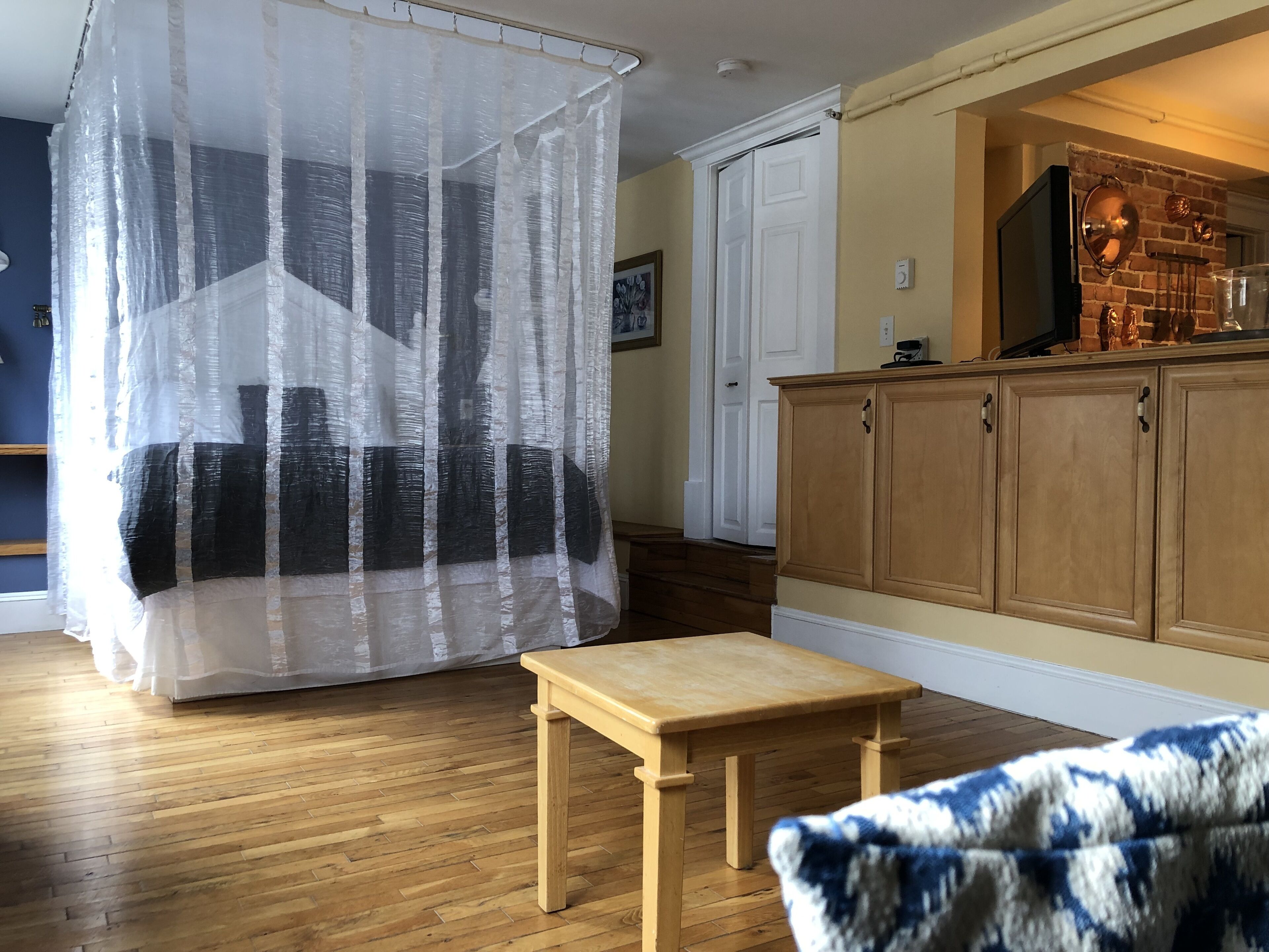 A room with a bed covered by a sheer curtain, a wooden table, a chair, and a kitchen area with wooden cabinets and a brick wall.