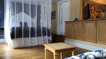 A room with a bed covered by a sheer curtain, a wooden table, a chair, and a kitchen area with wooden cabinets and a brick wall.