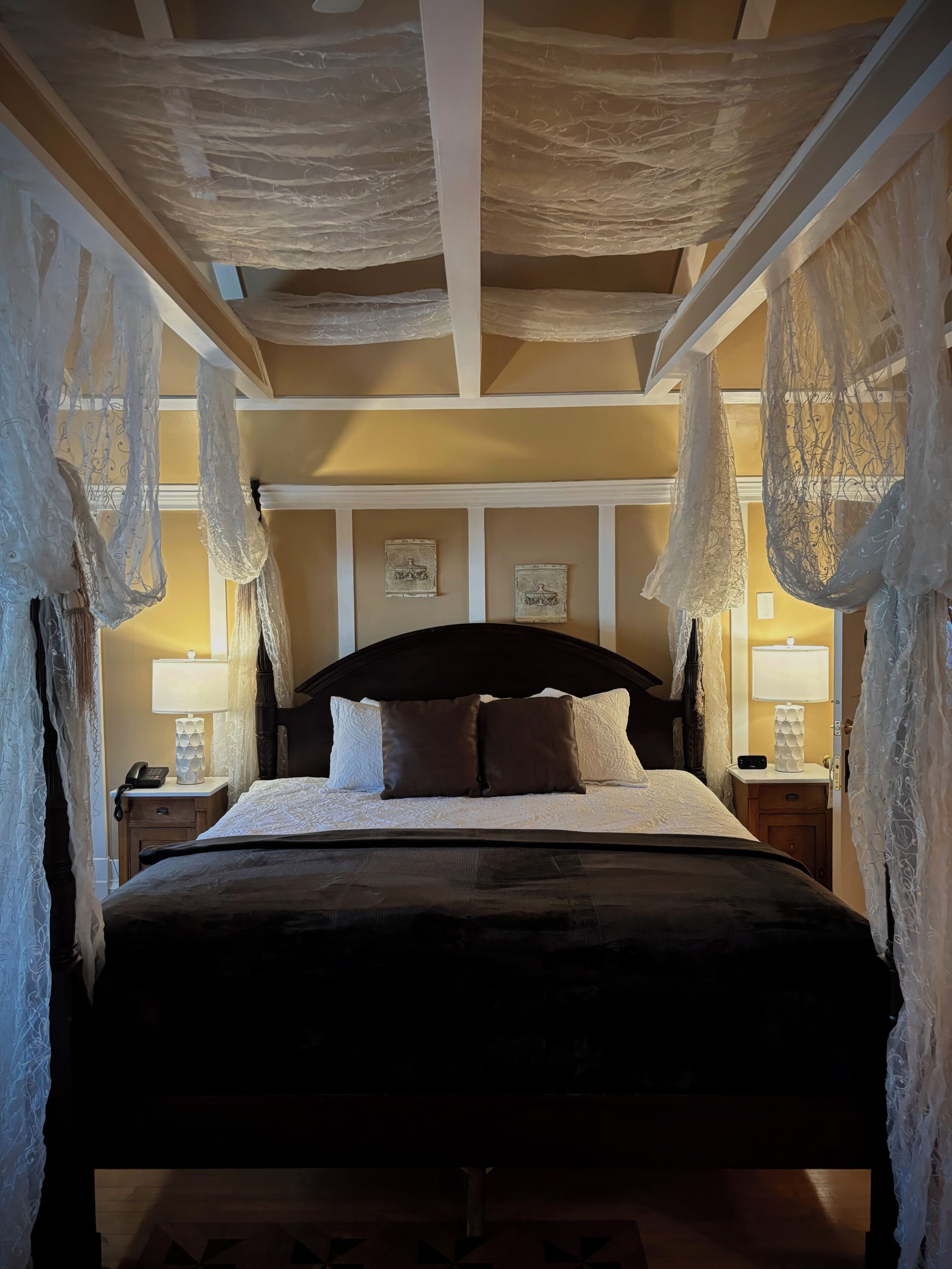 Una cama con cabecera oscura, ropa de cama blanca y dos almohadas marrones. Hay dos mesitas de noche con lámparas y un teléfono. La habitación tiene un techo de madera con vigas a la vista y paneles decorativos en las paredes.