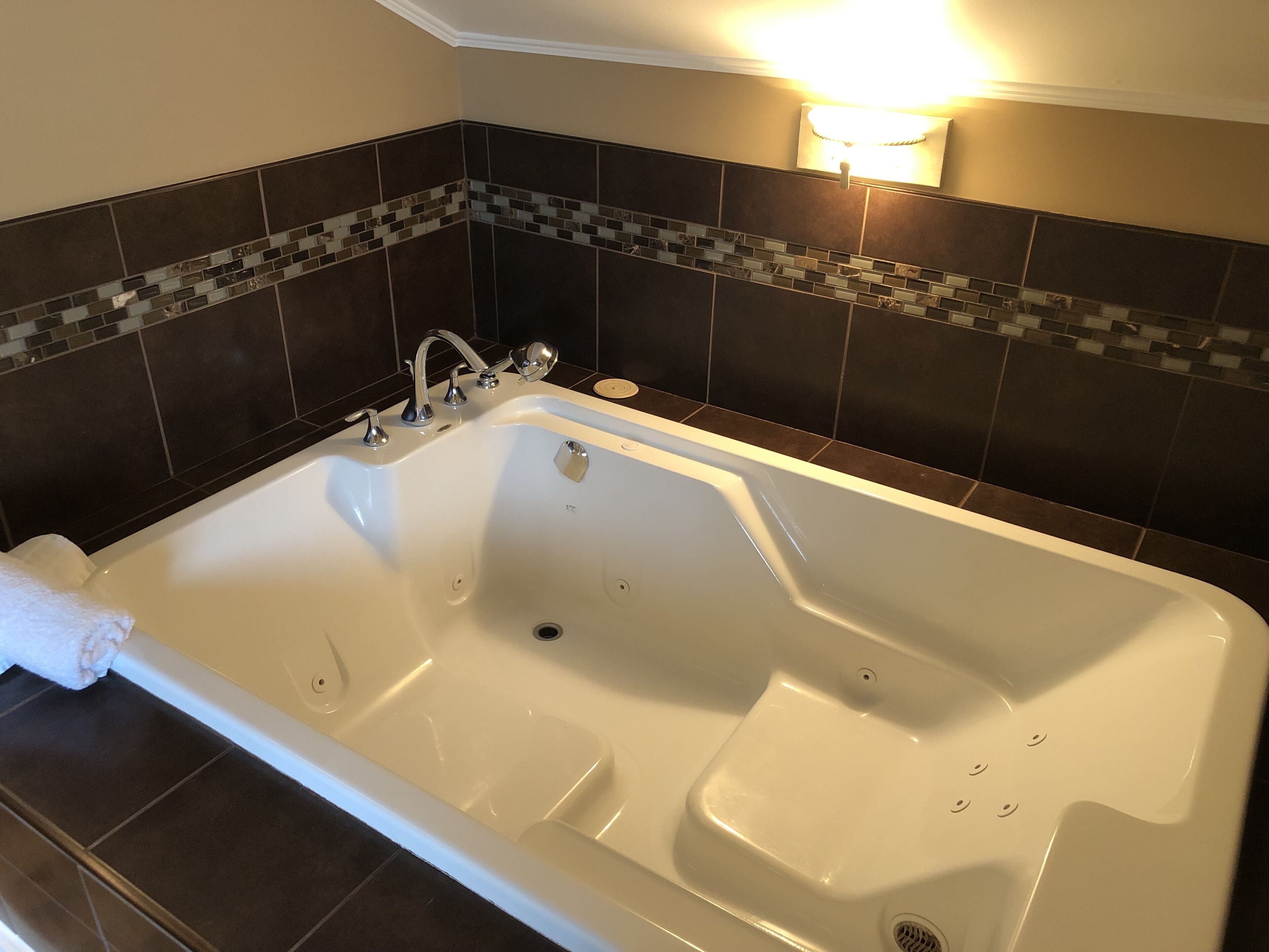 Une grande baignoire blanche, dotée d’un seul robinet et d’une douchette à main, est placée devant un mur recouvert de carreaux sombres, agrémenté d’une bordure en mosaïque.