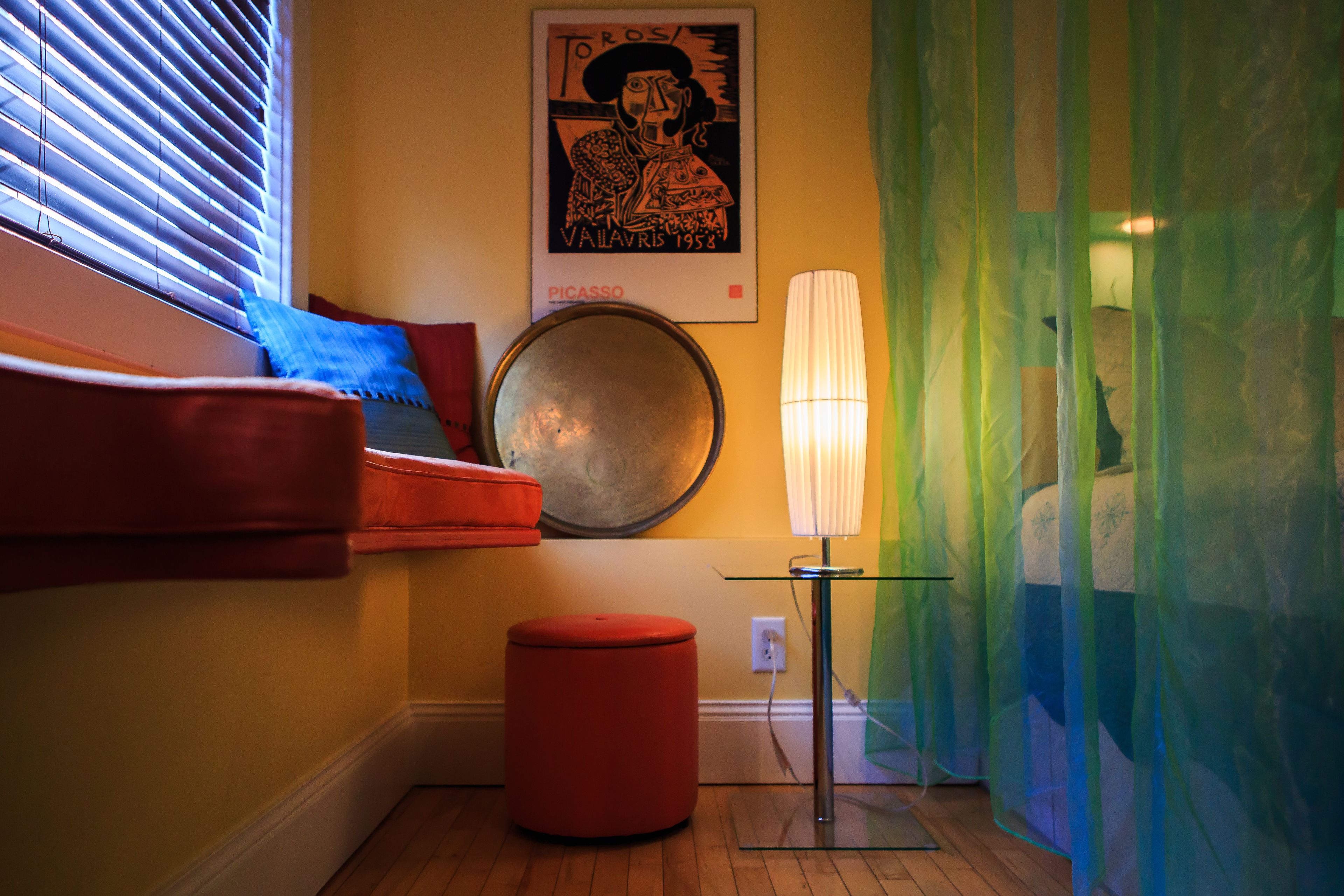 A room with a bed, a red ottoman, a wall-mounted lamp, a framed artwork, and a window with blinds.