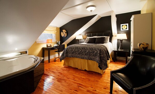 A bedroom with a large bed, a sitting area with a chair, a desk, a lamp, and a bathroom with a bathtub. - Leaside Manor by The Leaside Group (St. John's)
