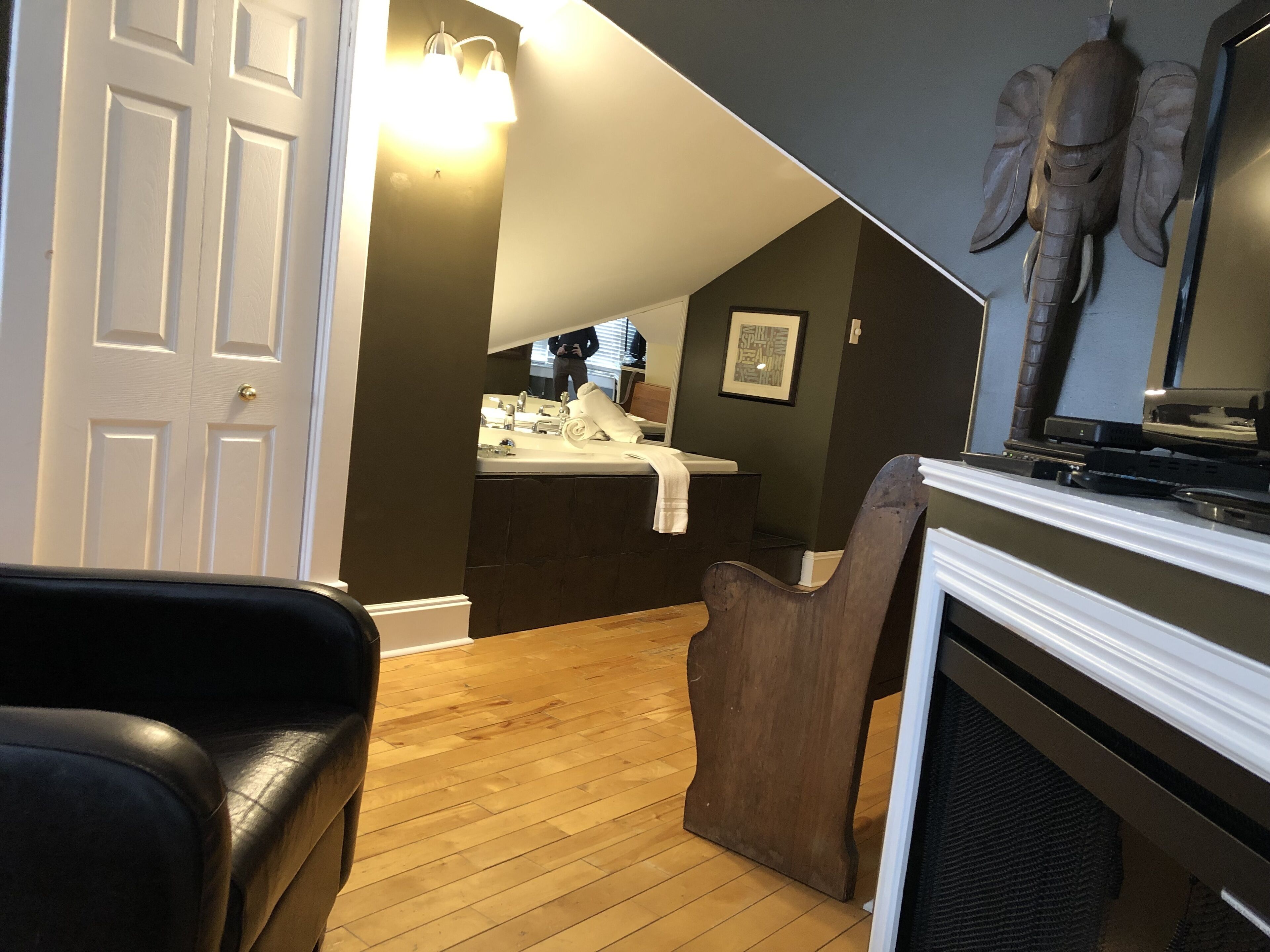 A room with a wooden floor, a white door, a dark brown wall, a white fireplace, and a wooden chair.