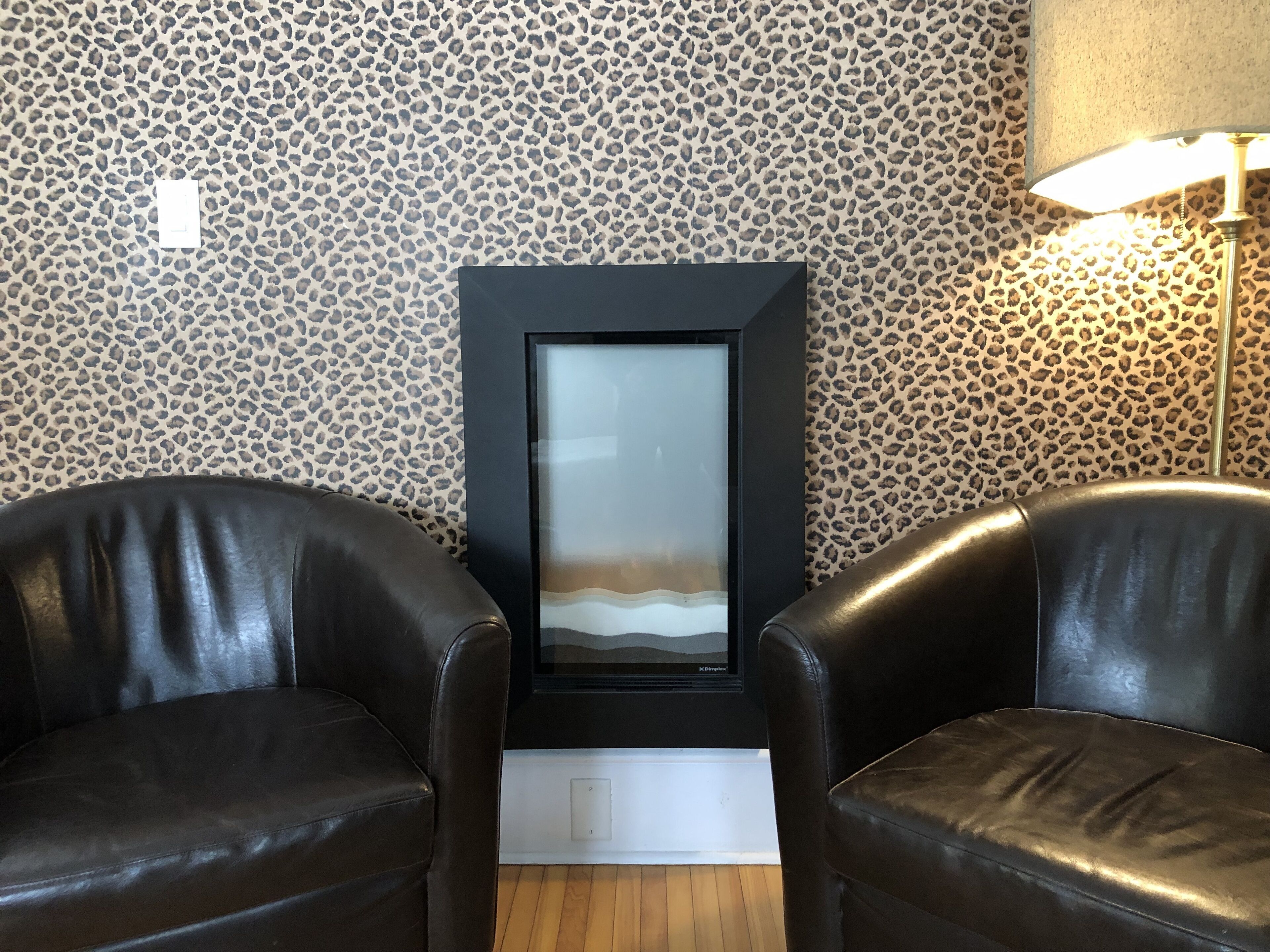 A room with leopard print wallpaper, two leather chairs, a framed artwork, and a floor lamp.
