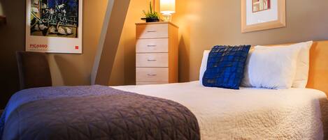 A hotel room with a bed, a dresser, a lamp, and two framed pictures on the wall.