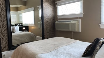 Een slaapkamer met een grote spiegel, een bed met een witte quilt, een kussen met luipaardprint en een airconditioning aan de muur.