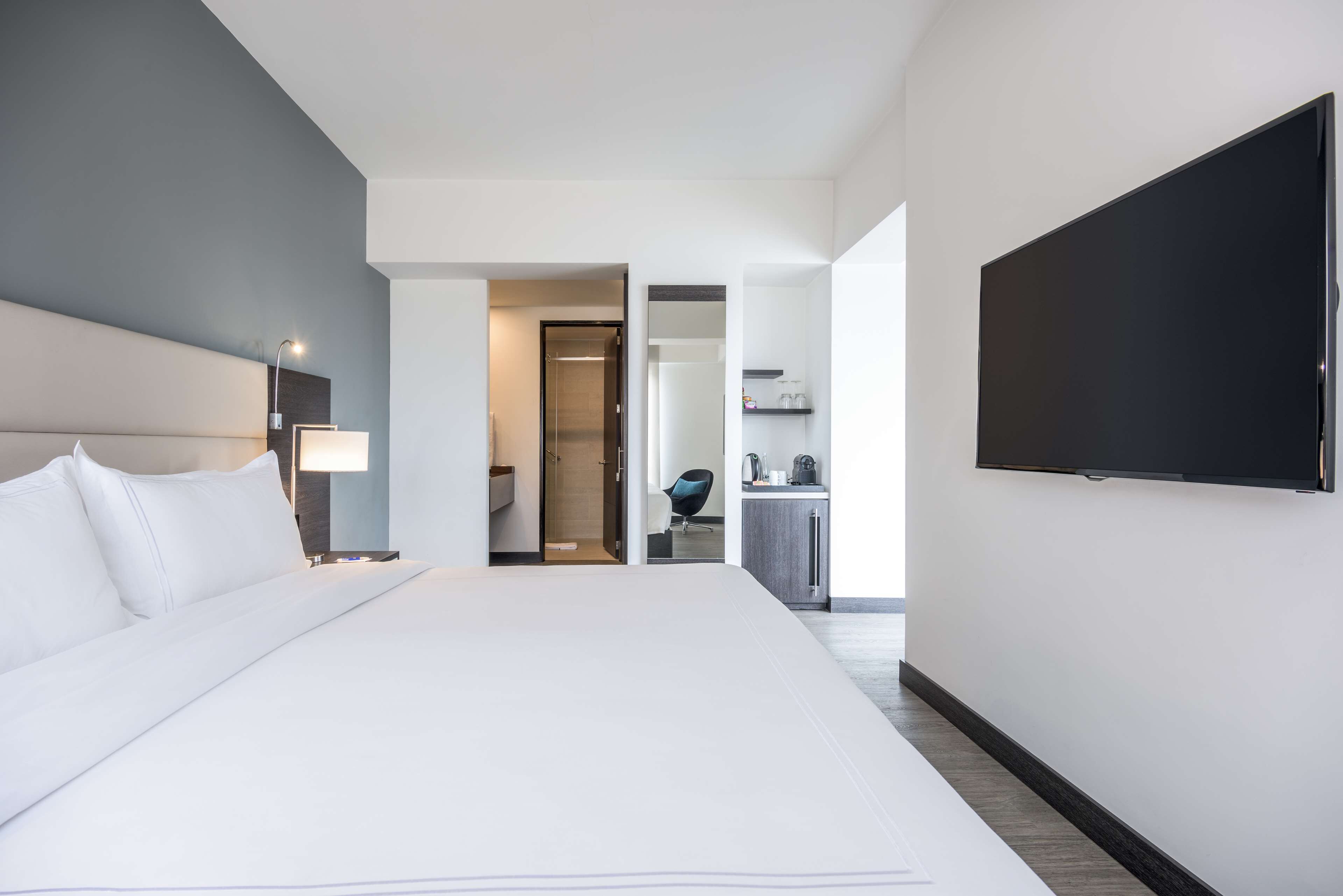 A modern hotel room with a large bed, a flat-screen TV, a desk with a chair, and a kitchenette.