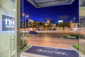 Indgangen til NH Hotels med en velkomst måtte, en parkeret bil udenfor og et gadesyn med bygninger og træer.