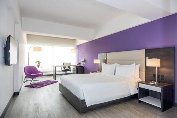 A modern hotel room with a large bed, a desk, a purple chair, and a TV on the wall.