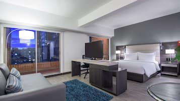 A modern hotel room with a large bed, a desk with a TV, a sofa, and a view of the city at night.