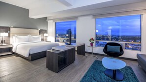 Habitación de hotel moderna con una cama grande, un escritorio, una silla y vistas a la ciudad al atardecer.