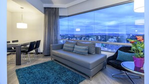 Una sala de estar moderna con un sofá gris, una mesa de comedor con sillas, un ventanal con vista a la ciudad y una alfombra azul.