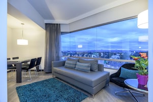 Una sala de estar moderna con un sofá gris, una mesa de comedor con sillas, un ventanal con vista a la ciudad y una alfombra azul.
