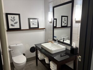 A modern bathroom with a white toilet, a white sink with a black countertop, and a large mirror. There are framed leaf art pieces on the wall above the sink.