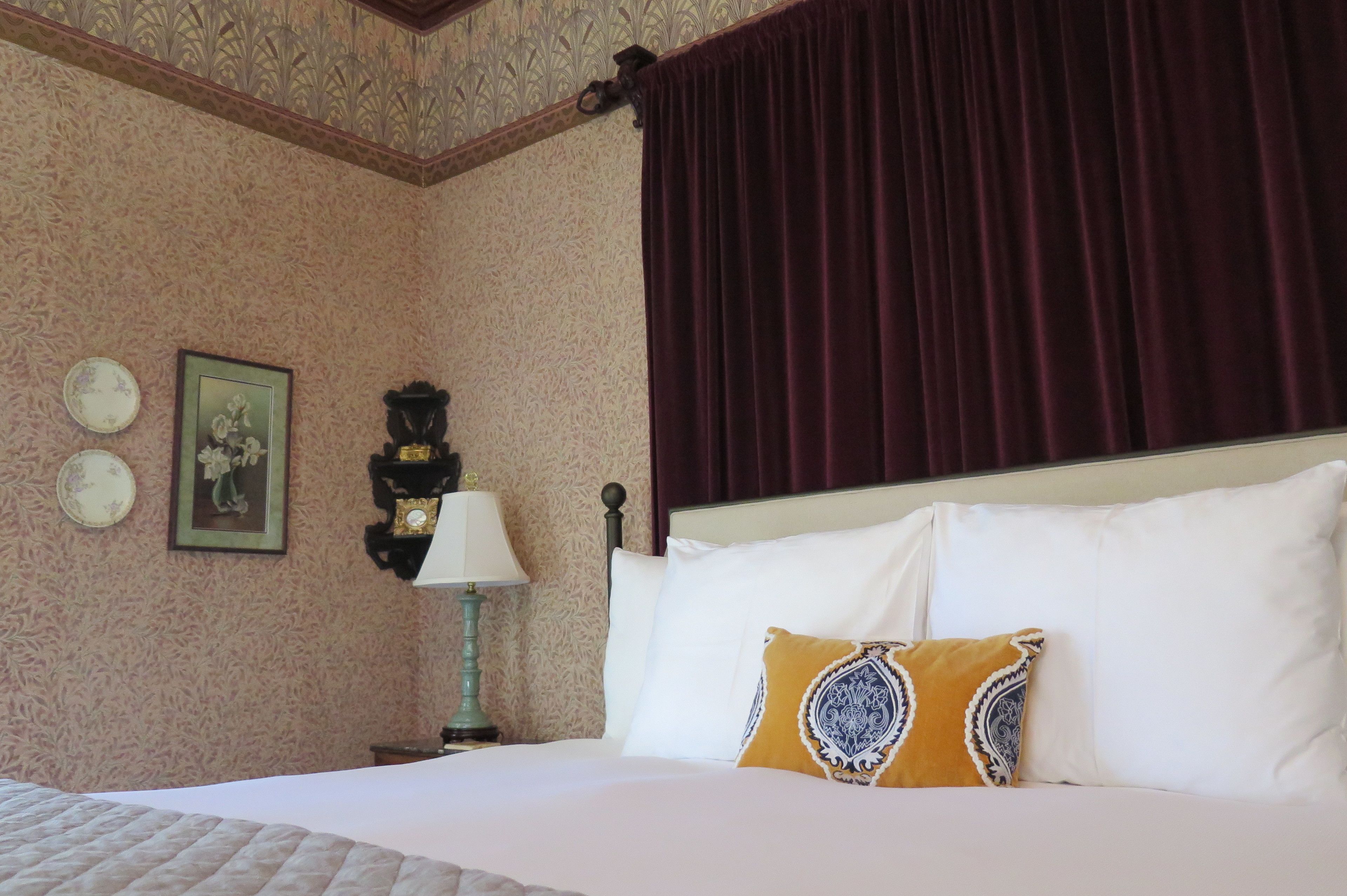 Una habitación de hotel con una cama, una mesita de noche con lámpara, un reloj de pared y un cuadro enmarcado.