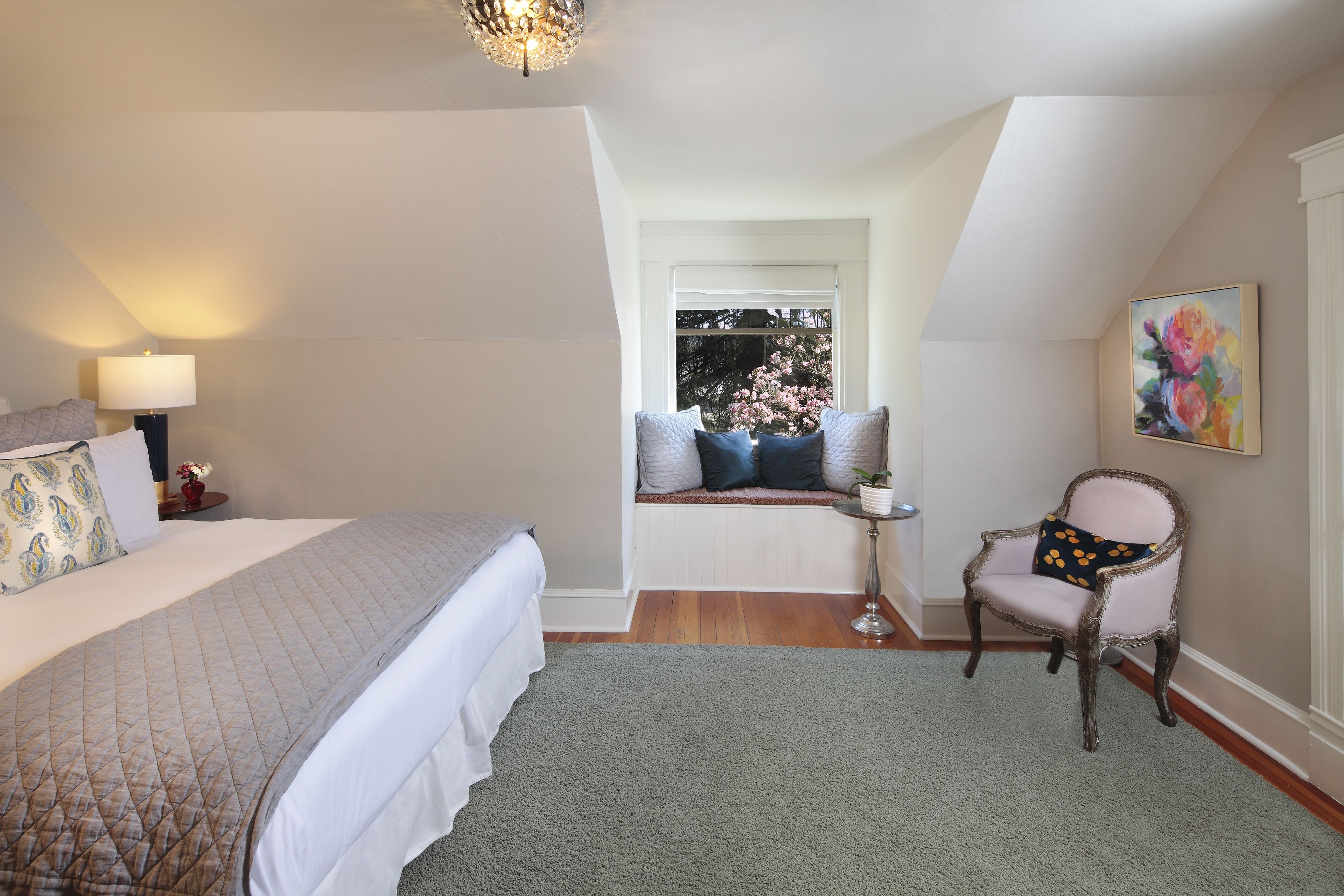 Quarto com uma cama grande, uma cadeira, uma mesinha e janela com vista para árvores.