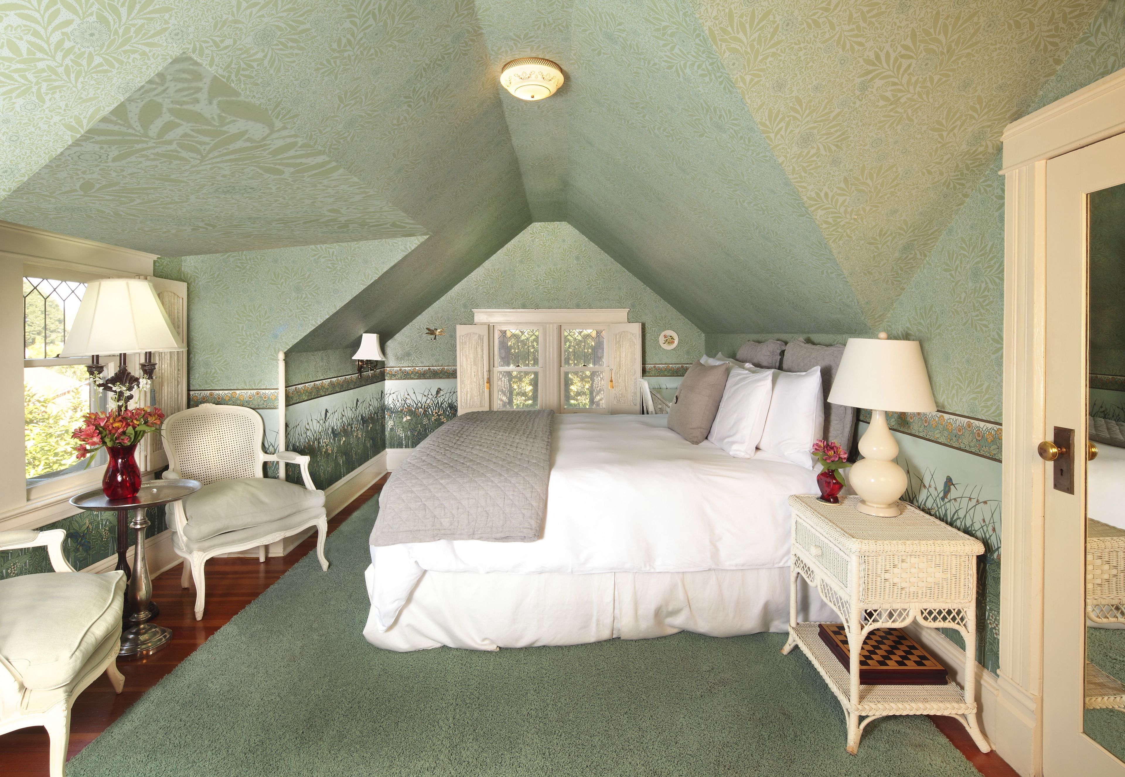 A bedroom with a high ceiling, a bed, a chair, a nightstand, and a window with a view.