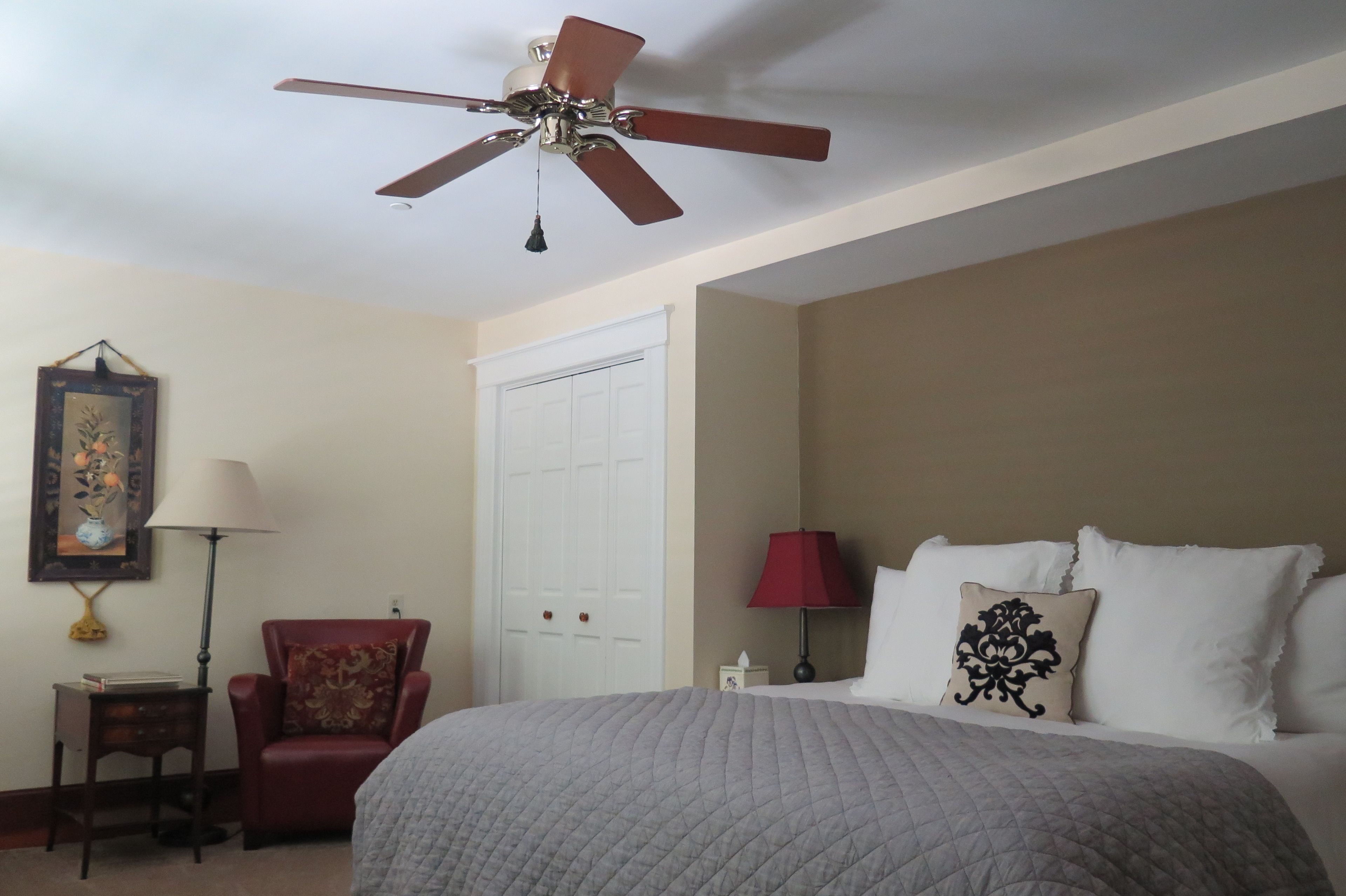Une chambre d’hôtel avec un lit, un fauteuil rouge, un ventilateur de plafond, une porte blanche et un tableau encadré au mur.