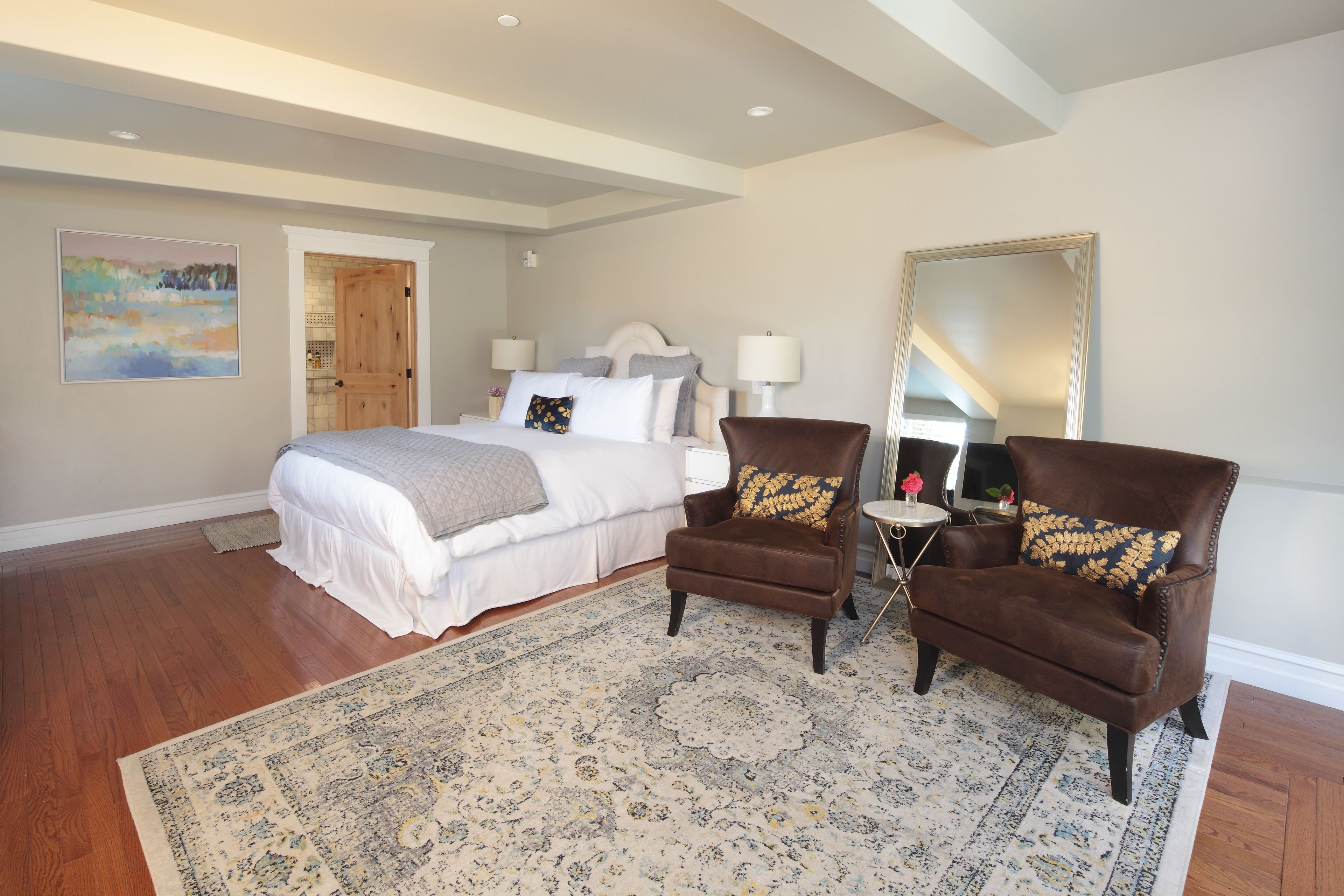 A bedroom with a large bed, two armchairs, a rug, a mirror, and a door leading to another room.