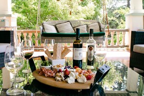A table set with a cheese board, wine glasses, and bottles, with a green sofa and lush greenery in the background.