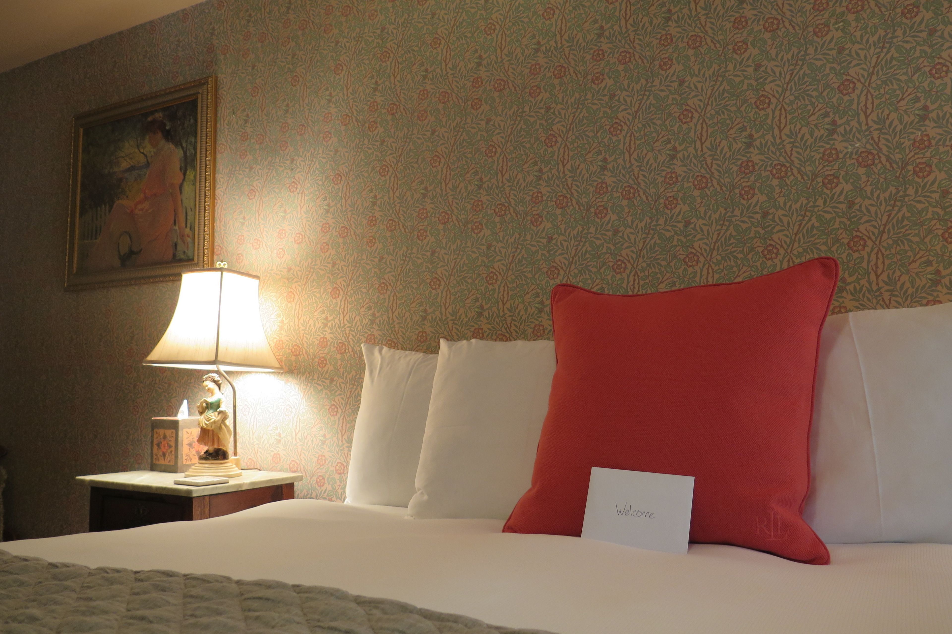 A hotel room with a bed, a nightstand with a lamp, a painting on the wall, and a welcome card on the bed.